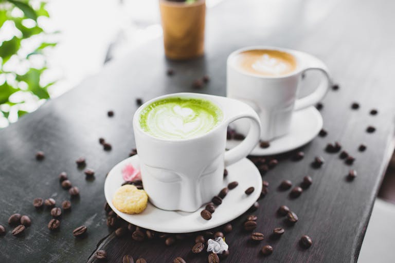 From above of refreshing tasty matcha green tea latte and cappuccino in creative face shaped mugs placed on wooden table with scattered coffee beans